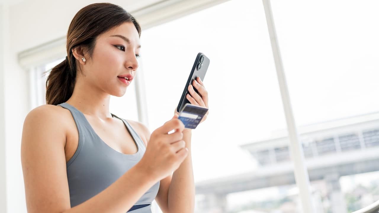 Fit young woman wearing workout gear holds a smartphone and credit card while on a treadmill.