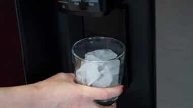 A woman stands in front of a sleek, stainless steel refrigerator, holding a glass filled to the brim with perfectly cubed ice