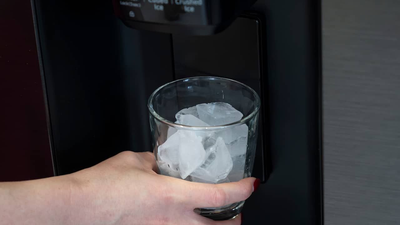 A woman stands in front of a sleek, stainless steel refrigerator, holding a glass filled to the brim with perfectly cubed ice