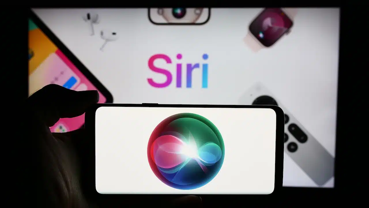 Person holding cellphone with logo of digital assistant software Siri (Apple Inc.) in front of business webpage. Focus on phone display.