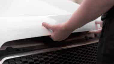 hands in black casual clothes opening the hood of a white car.