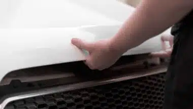 hands in black casual clothes opening the hood of a white car.