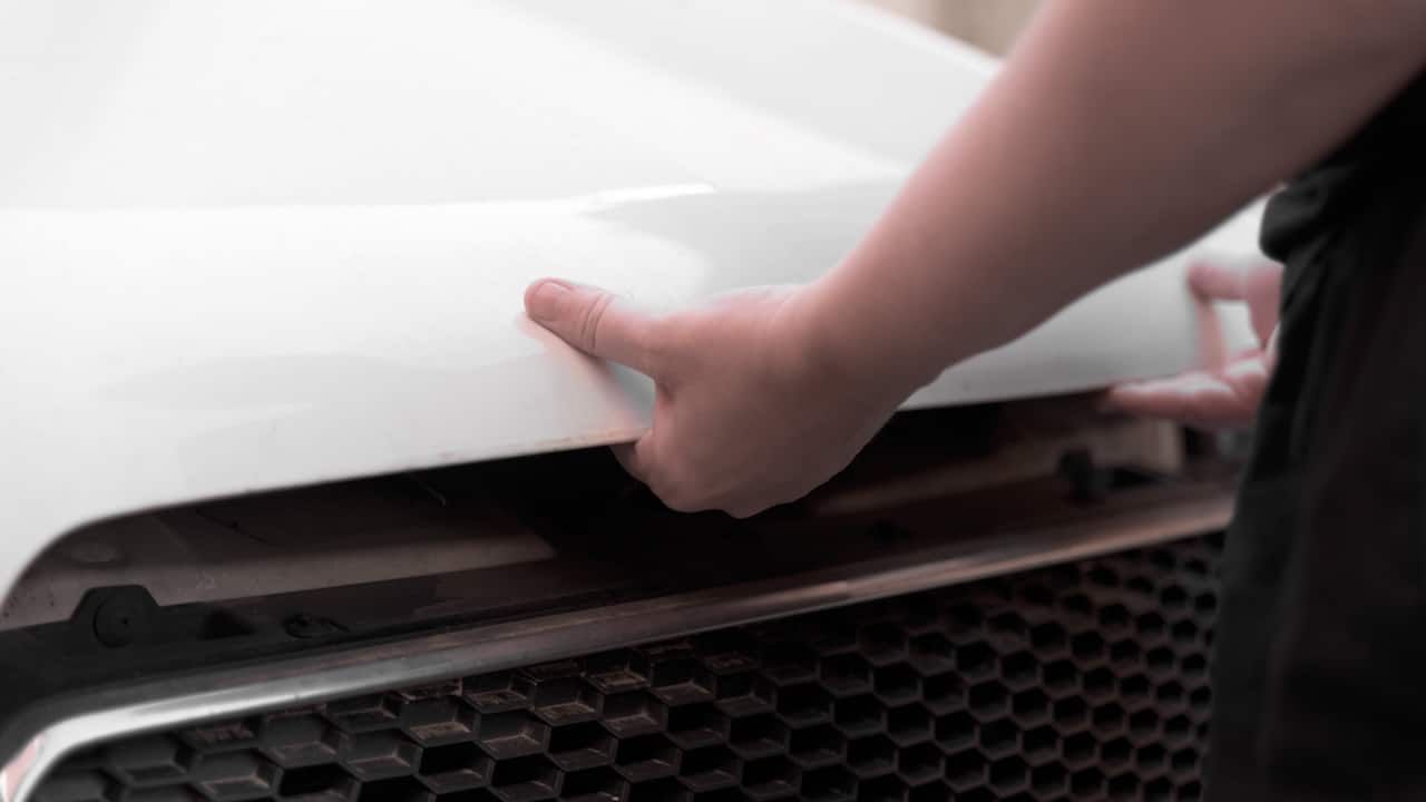 hands in black casual clothes opening the hood of a white car.