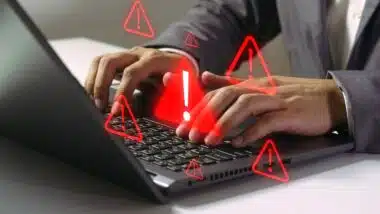 Close up of hands typing on a laptop with multiple red glowing caution symbols overlaying the keyboard representing a security breach.