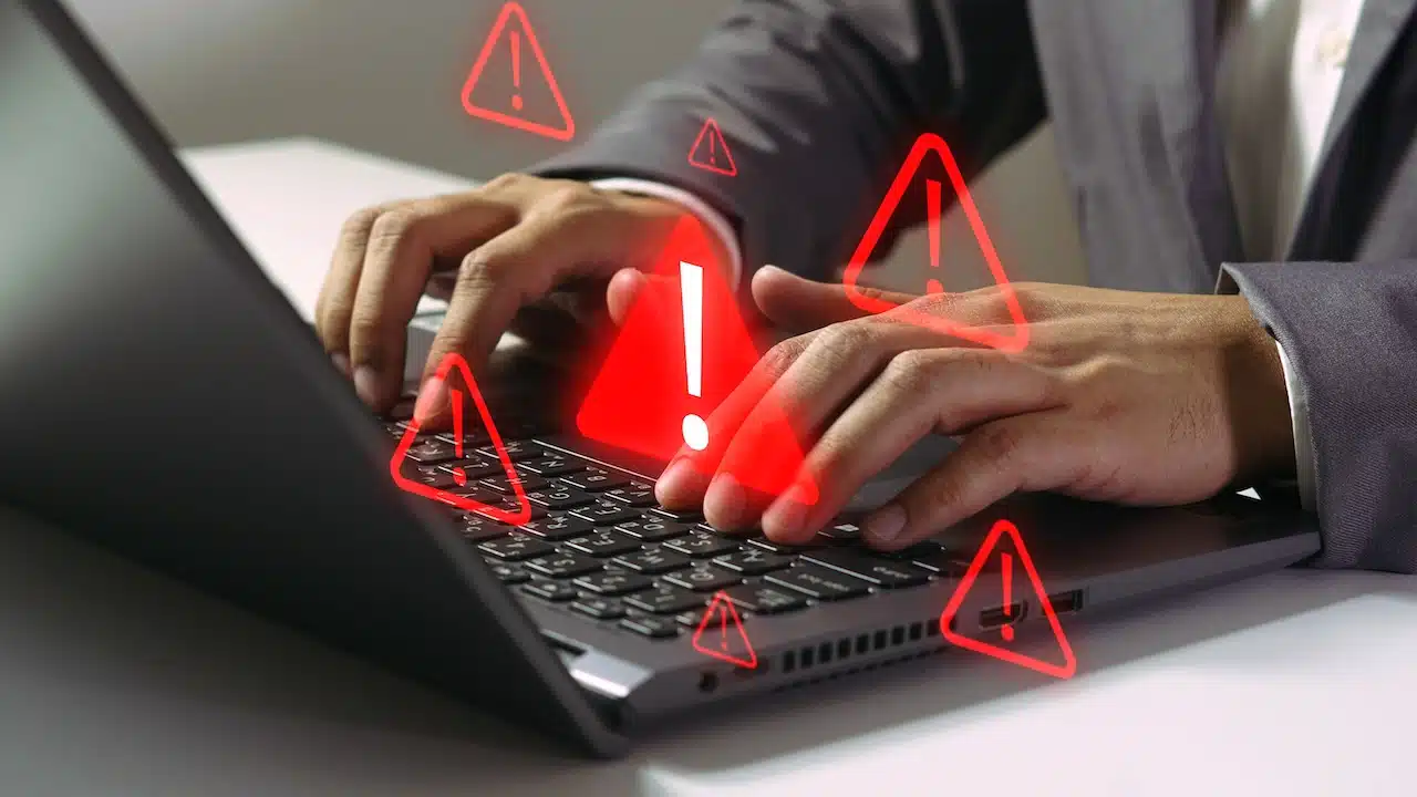 Close up of hands typing on a laptop with multiple red glowing caution symbols overlaying the keyboard representing a security breach.