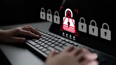 A person typing on a keyboard with a warning symbol on the screen signaling a data breach.