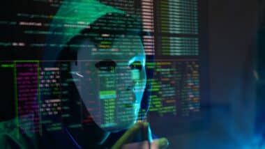 male hacker wearing a white mask and a hoodie sits in front of a screen,