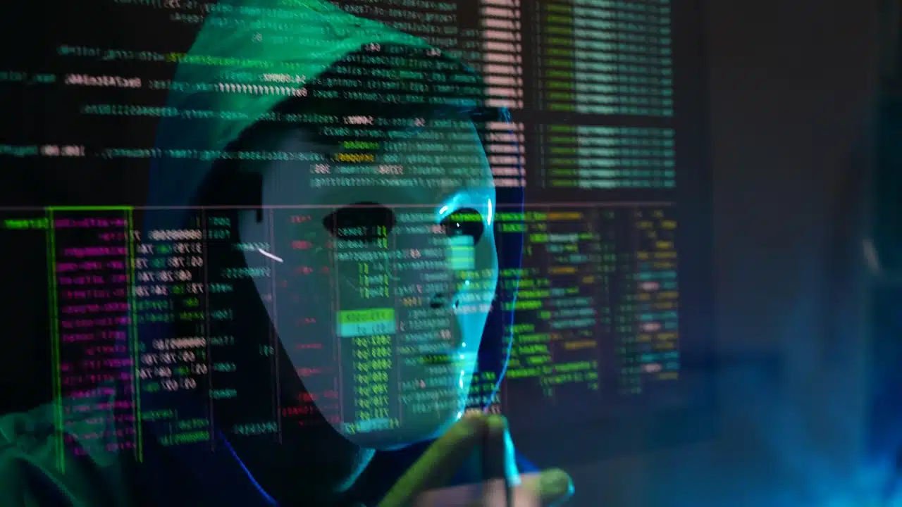 male hacker wearing a white mask and a hoodie sits in front of a screen,