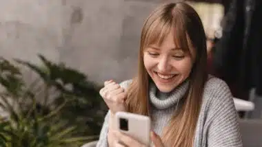 Joyful blonde woman playing video game on mobile phone, reading good news, celebrating winning lottery, feeling so happy and cheerful while making winner gesture, sitting at table.
