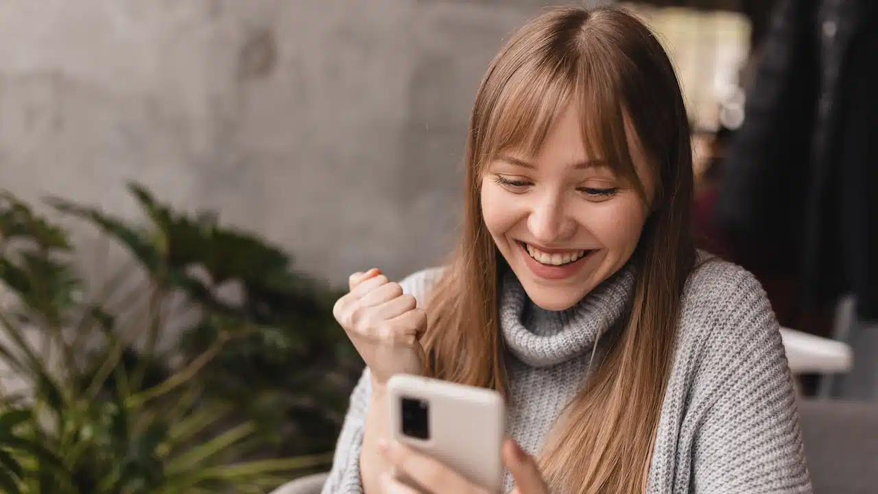 Joyful blonde woman playing video game on mobile phone, reading good news, celebrating winning lottery, feeling so happy and cheerful while making winner gesture, sitting at table.
