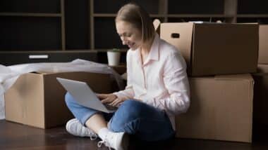 Woman sit on floor with cardboard boxes use laptop make order, buy, furniture.