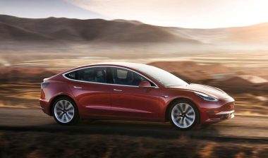 A red Tesla model 3 driving along a roadway, representing the Tesla recall.