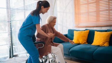 Assistant helping elderly woman onto her couch.