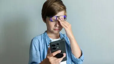 A young boy rubbing his eyes while using a smartphone, representing social media warning labels.