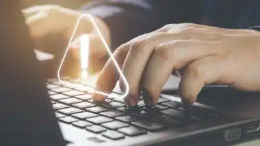 Close up of hands typing on a laptop with a warning symbol overlay, representing the PowerSchool data breach.