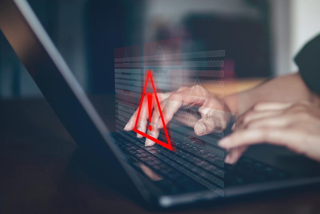 Close up of hands typing on a laptop with a warning symbol overlay, representing the American Addiction Centers data breach.