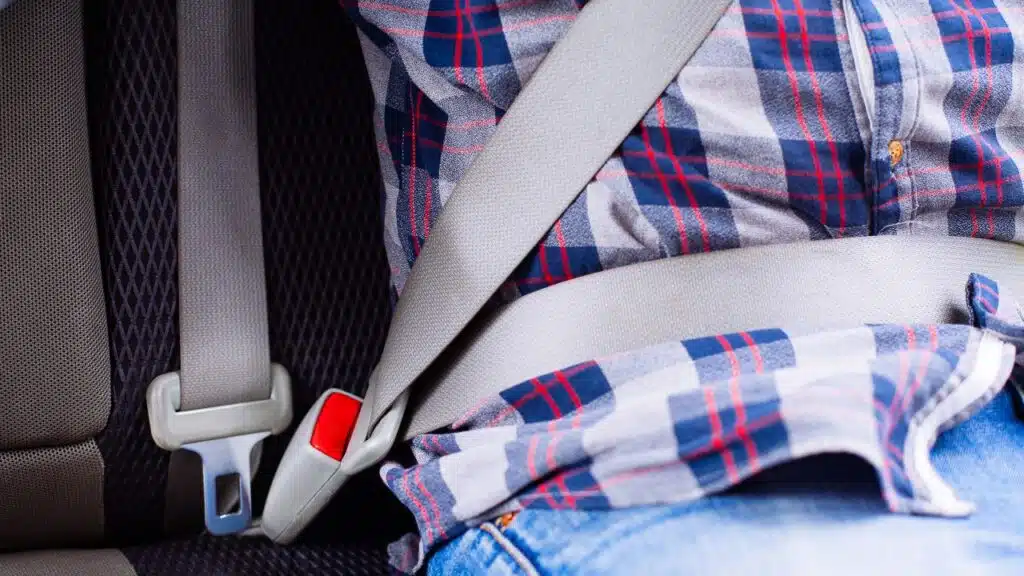 Photo of man in plaid shirt sitting in a car putting on seat belt. 