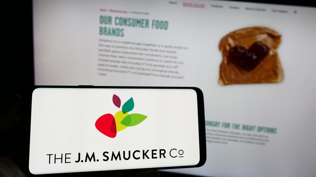 Person holding cellphone with logo of US food business The J.M. Smucker Company on screen in front of business webpage.