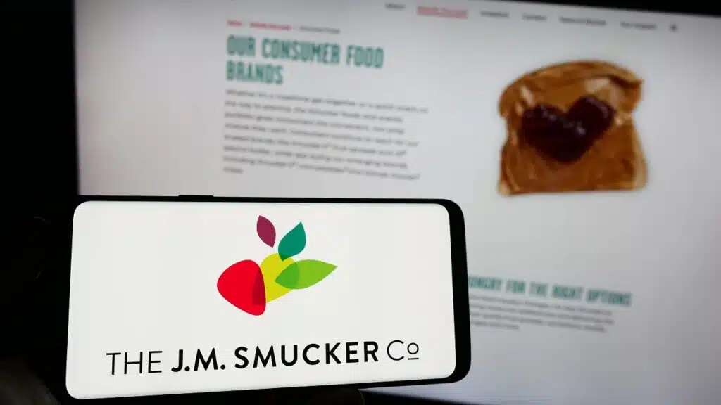 Person holding cellphone with logo of US food business The J.M. Smucker Company on screen in front of business webpage.