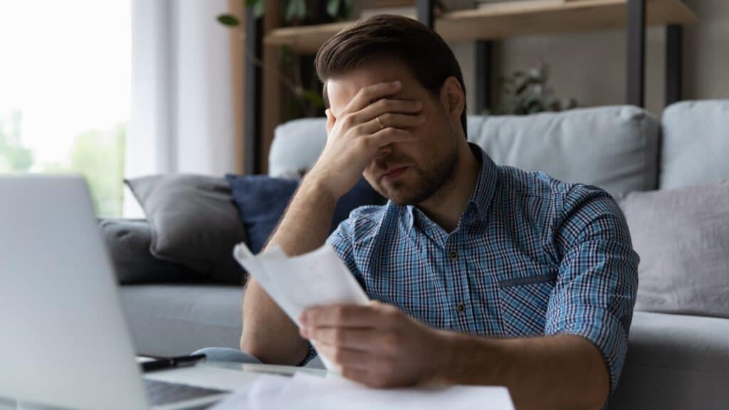 Frustrated desperate millennial man checking bills for payments, holding receipts, high mortgage fees.