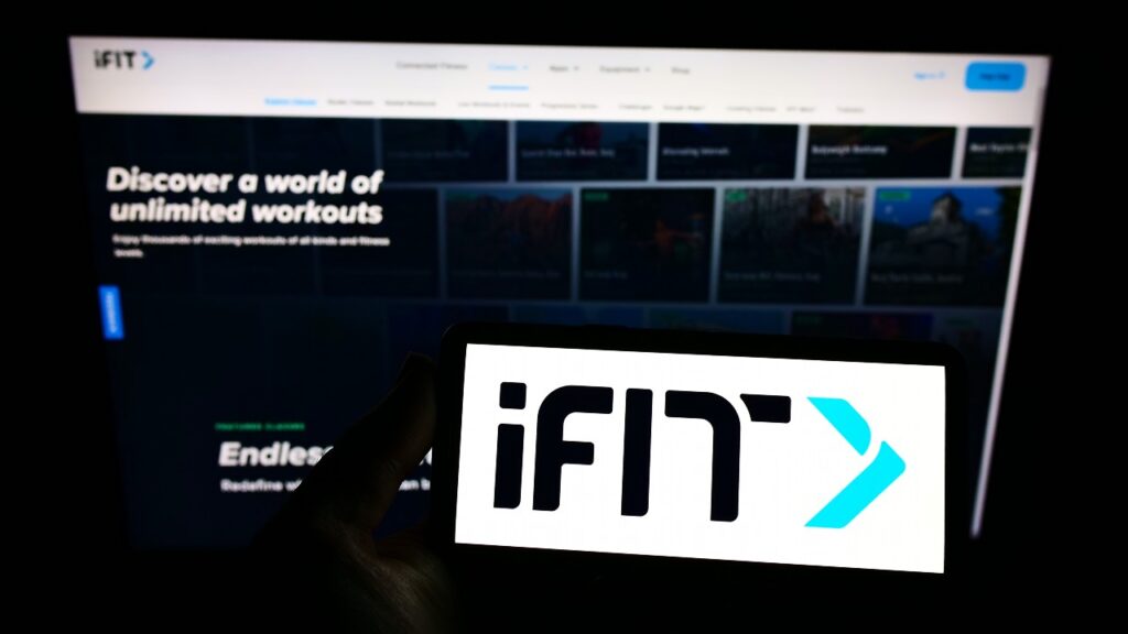 Person holding smartphone with logo of US fitness company iFIT Inc. on screen in front of website.