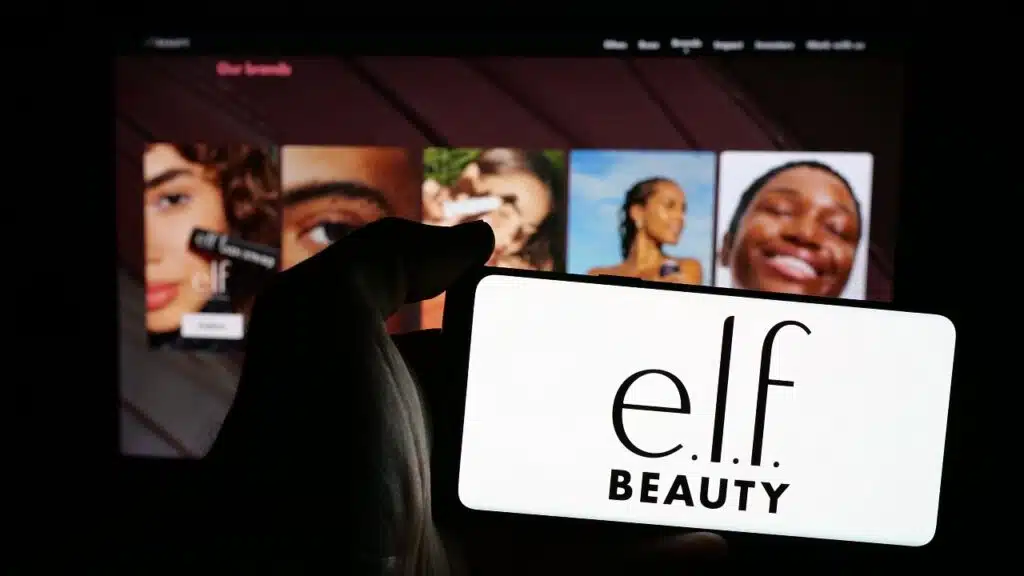 Person holding mobile phone with logo of American cosmetics company e.l.f. Beauty Inc. on screen in front of web page