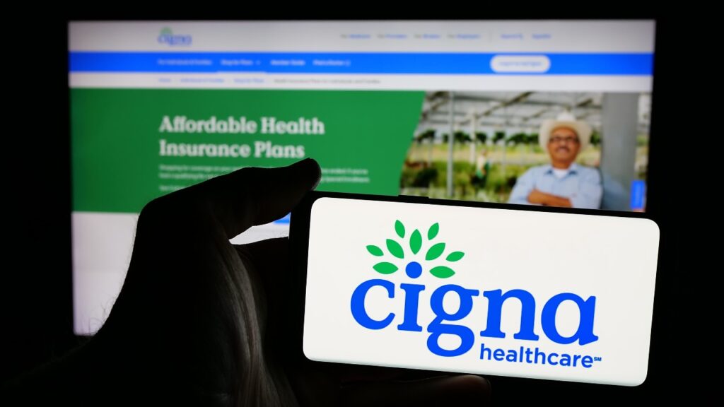 Person holding mobile phone with logo of American health company company Cigna Healthcare on screen in front of web page.