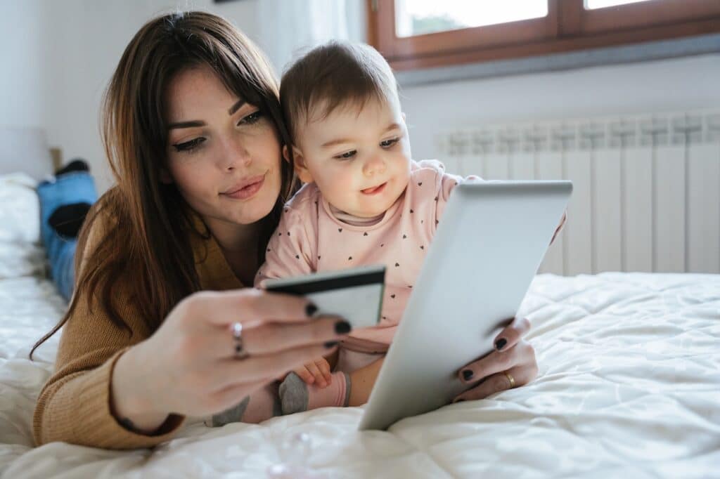 Portrait of a beautiful young mom with a baby girl lying on the bed in a moment of intimacy while buying products online with the tablet and holding her credit card in hand.