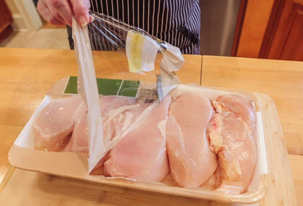 Female chef opening package of fresh boneless chicken breasts. Raw chicken breast in grocery store packaging on a wooden kitchen table.