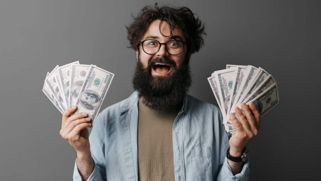A joyous bearded man with glasses celebrates financial success, holding several U.S. dollar bills in both hands against a neutral background.