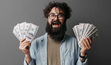 A joyous bearded man with glasses celebrates financial success, holding several U.S. dollar bills in both hands against a neutral background.