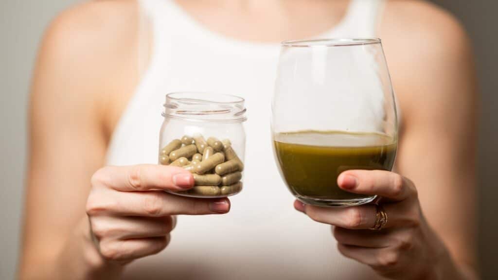 Kratom consumption concept: female holds a ready to drink glass of green liquid, powdered kratom melted in hot water, glass of tea and handful of pills in the jar, close up photo, selective focus
