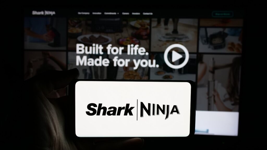 Person holding cellphone with logo of US technology company SharkNinja on screen in front of business webpage.