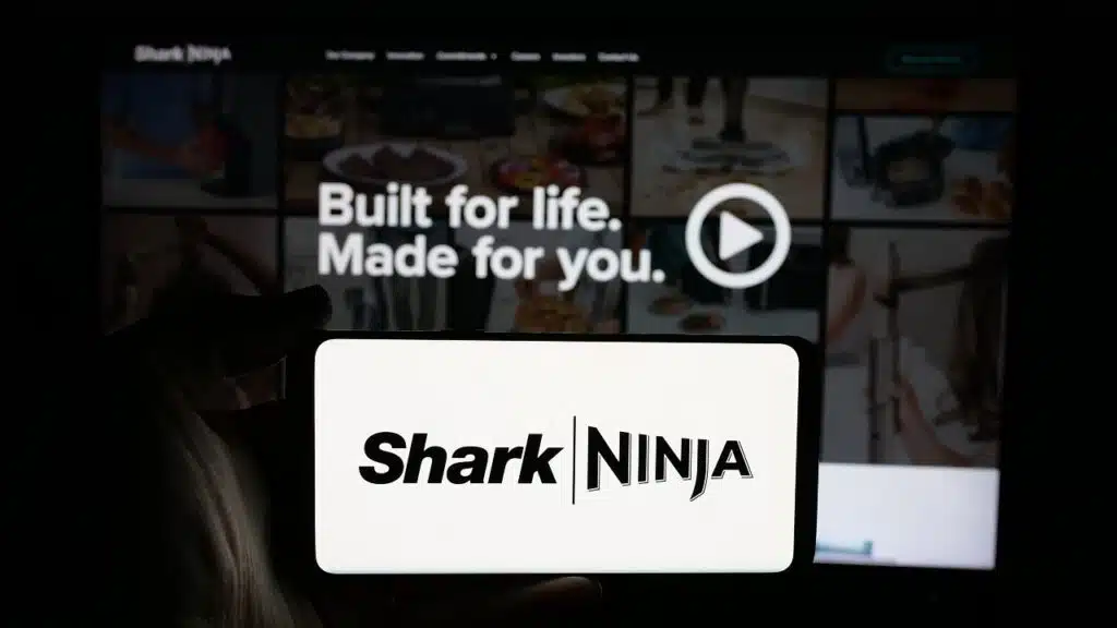 Person holding cellphone with logo of US technology company SharkNinja on screen in front of business webpage.