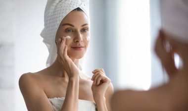 Attractive young adult woman apply facial cream look in mirror, beautiful healthy lady wrapped in towels put moisturizing lifting nourishing day creme on soft hydrated moisturized skin in bathroom