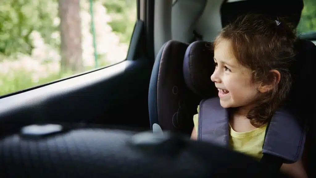 Safe movement of children in the car. Adorable baby girl enjoys the travel by car, looks out of an opened window while sitting in a safety booster car seat
