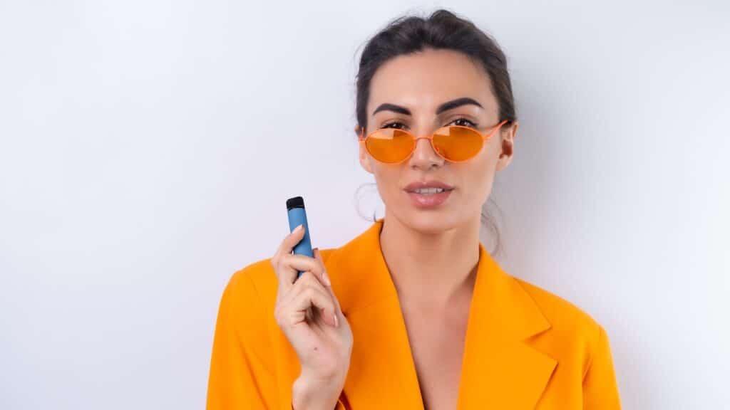 A young woman in trendy stylish glasses and a bright orange oversized jacket on a white background smokes an electronic cigarette