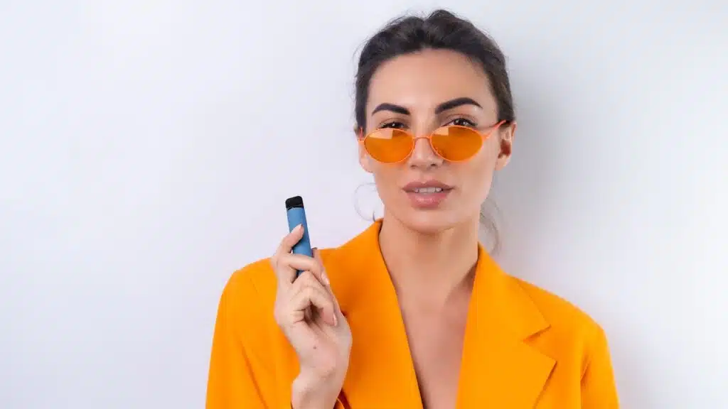 A young woman in trendy stylish glasses and a bright orange oversized jacket on a white background smokes an electronic cigarette