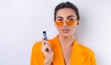 A young woman in trendy stylish glasses and a bright orange oversized jacket on a white background smokes an electronic cigarette