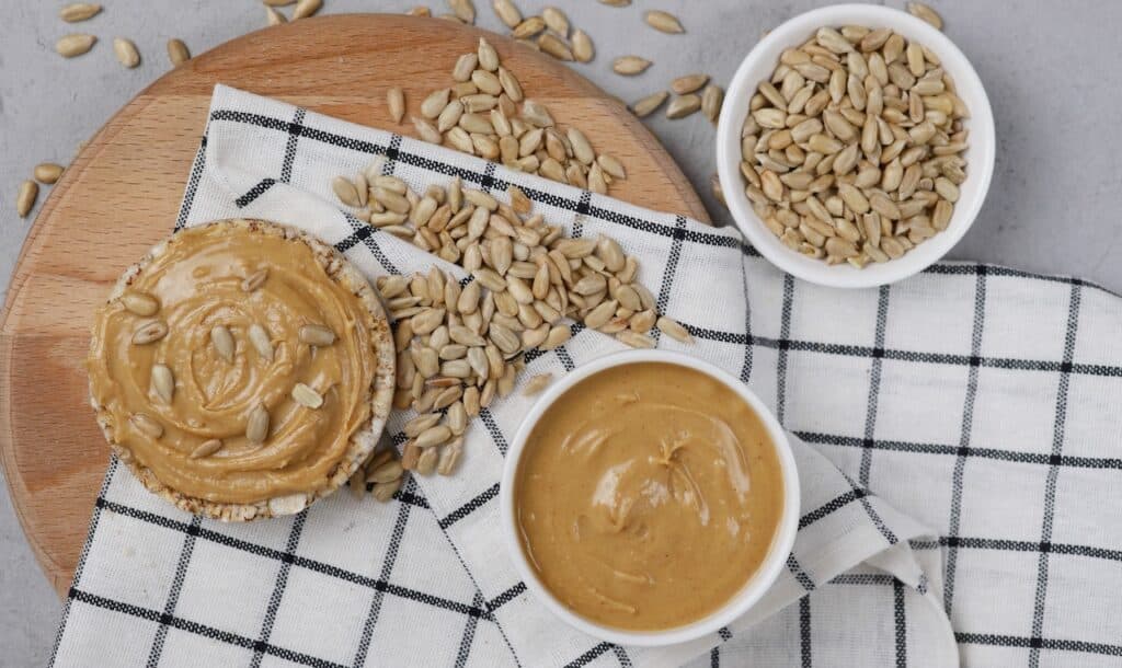 Organic sunflower butter, sunflower seeds in white bowls with wheat bread and scattered sunflower seeds on a gray background.