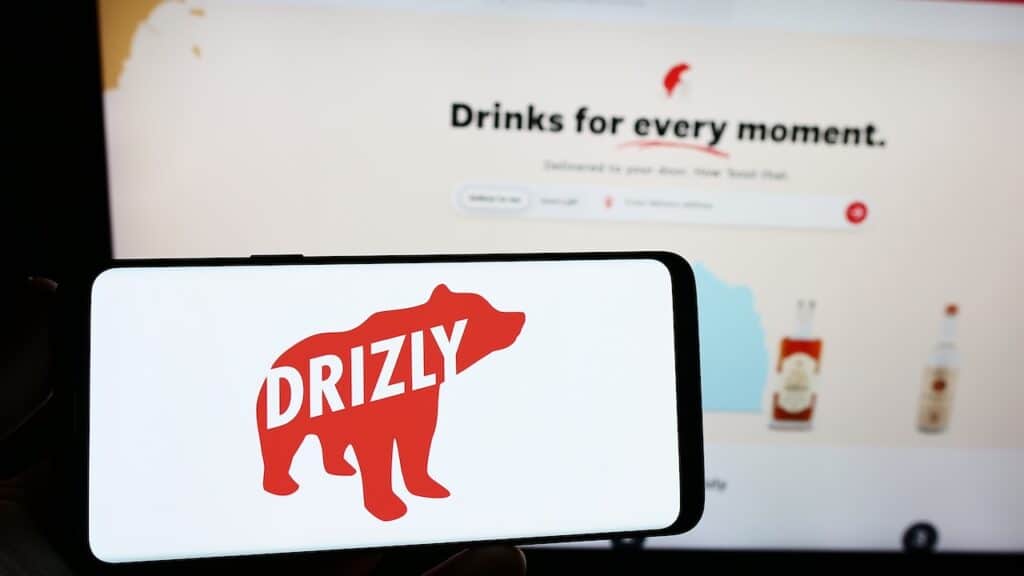 Person holding smartphone with logo of US alcohol delivery company Drizly LLC on screen in front of website. Focus on phone display.