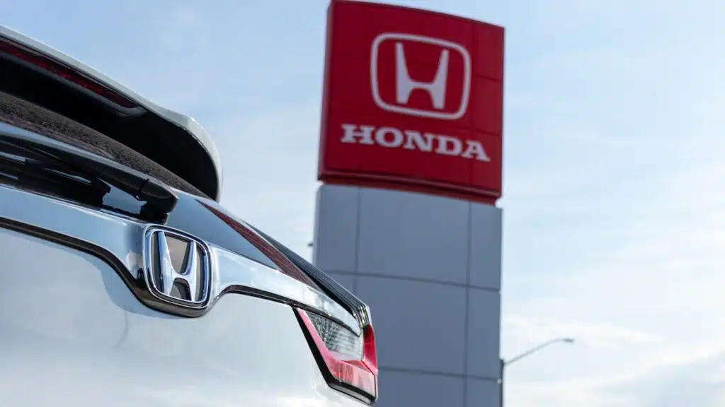 Honda emblem on a car at a dealership, with the Honda Motor Company logo on a sign in the background.