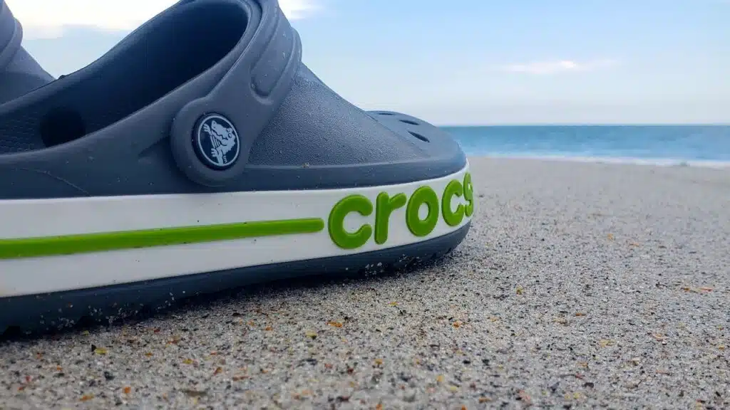 Gray Crocs sandals lie on the sand by the ocean. trendy croc beach shoes.