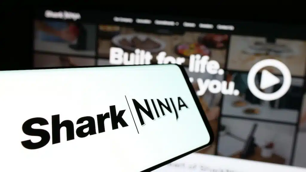 Mobile phone with logo of American technology company SharkNinja on screen in front of business website. Focus on center of phone display.
