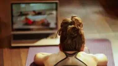 Seen from behind fit sports woman in fitness clothes in the modern living room using online fitness training program in laptop while laying on fitness mat.
