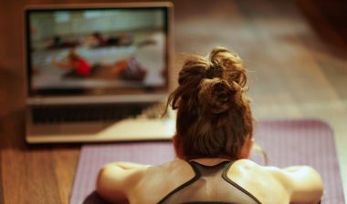 Seen from behind fit sports woman in fitness clothes in the modern living room using online fitness training program in laptop while laying on fitness mat.