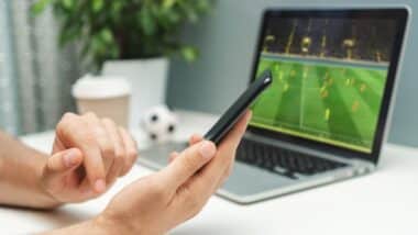 Close up cropped shot of male hand holding smartphone. Man watching soccer play live broadcast online on his laptop and making bets on his favourite team using mobile application.