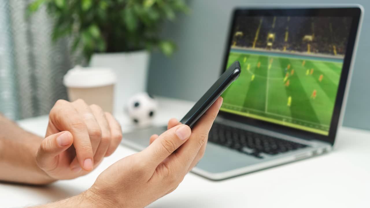 Close up cropped shot of male hand holding smartphone. Man watching soccer play live broadcast online on his laptop and making bets on his favourite team using mobile application.