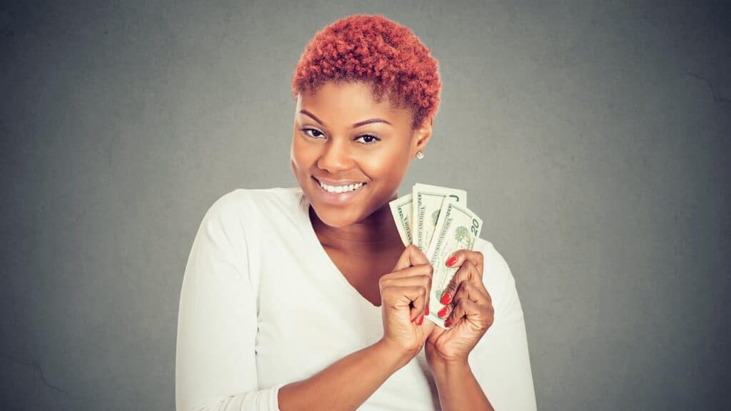 Portrait of super happy excited successful young woman holding money dollar bills in hand, isolated on gray background.