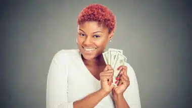 Portrait of super happy excited successful young woman holding money dollar bills in hand, isolated on gray background.